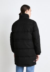 Svart pufferjakke med høy krage, med en kviltet design og lange ermer. Merkeplaten er tydelig nær kanten. Bært over blå jeans.
