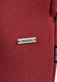 Červená tkanina s jemnou pletenou strukturou má kovovou etiketu s vyrytým nápisem "SMILODOX." Na boku je viditelná černá visačka.