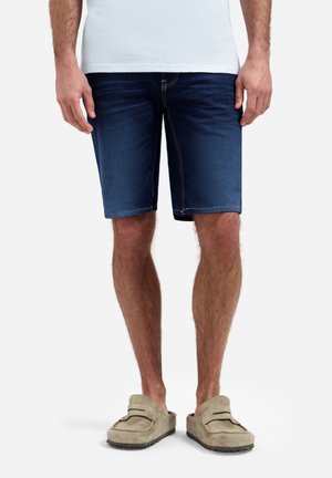 Man draagt donkerblauwe denimshorts, een wit overhemd en beige instappers, staand tegen een effen achtergrond.