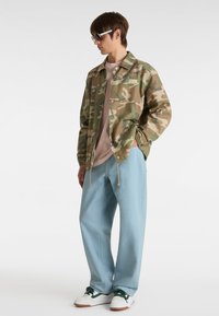 Camouflage jacket with a relaxed fit, paired with light blue pants and white sneakers. Features a logo patch on the jacket and drawstring details.