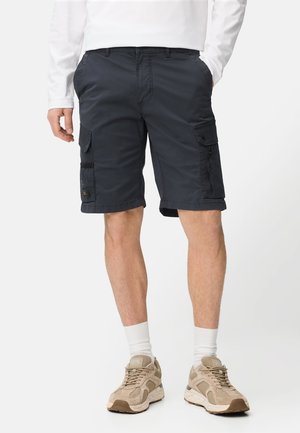 Mand iført sorte cargobukser, hvide sokker, beige sneakers og en hvid langærmet skjorte, med hænderne i lommerne, stående mod en hvid baggrund.