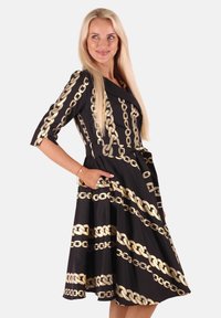 Blonde Frau in einem schwarzen Midi-Kleid mit goldenen Kettenmustern, steht mit einer Hand in der Tasche und blickt zur Seite, mit einem leichten Lächeln.