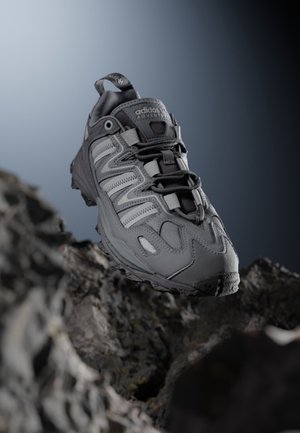 Grå Adidas vandresko med detaljerede syninger og tyk sål svævende over stenrig terræn mod en gradient grå-blå baggrund.