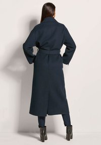 Lang marineblauwe jas van een gestructureerde wolmix, met een ceintuur in de taille, wijde mouwen en zijsplitten voor gemakkelijke beweging. Gekocht in combinatie met zwarte hoge laarzen.