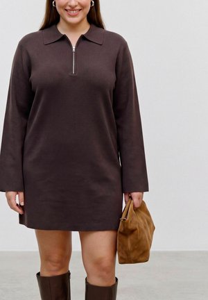 Femme portant une robe marron foncé à manches longues avec une fermeture éclair à l'avant, tenant une pochette en daim beige et portant des bottes hautes marron.
