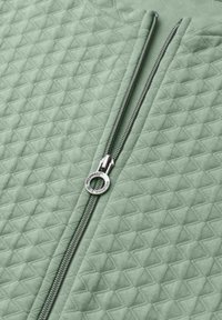 Tissu matelassé vert avec une fermeture éclair argentée partiellement ouverte, équipée d'une tirette ronde avec texte gravé.
