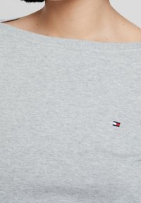 Pull gris en maille, présentant un col large et un petit logo brodé avec des éléments rouges, blancs et bleus sur la poitrine.