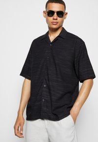 Black short-sleeve button-up shirt with a subtle textured stripe design and a spread collar. Model wears dark sunglasses.