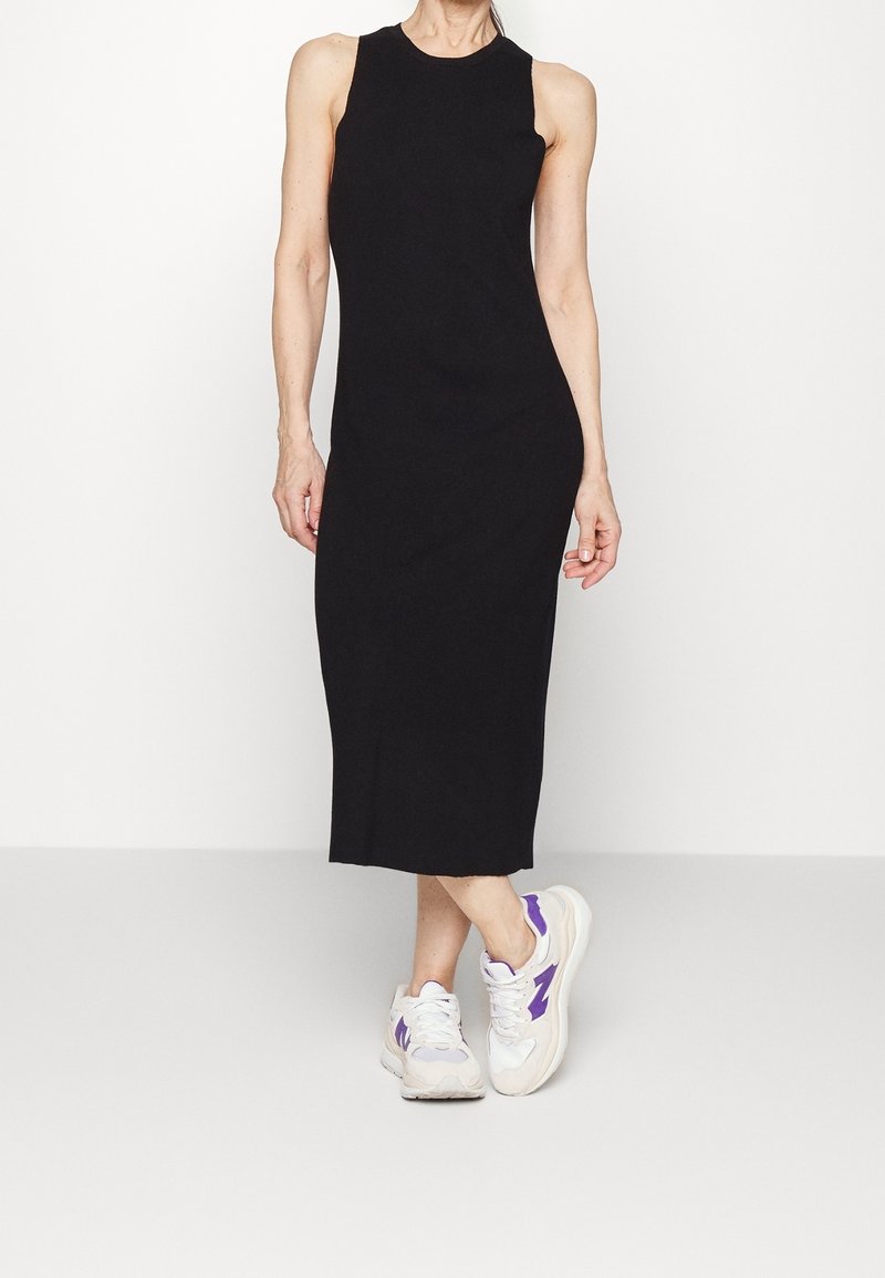 Femme portant une robe midi noire sans manches assortie à des baskets blanches et violettes, debout devant un fond blanc uni.