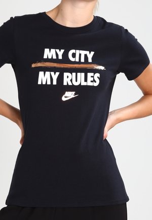 Czarna koszulka Nike z wyraźnym białym napisem "MY CITY" nad brązowym pociągnięciem pędzla i napisem "MY RULES" poniżej, noszona przez osobę z rękami na biodrach.