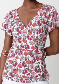 Vit omlottopp med blommönster, utrustad med korta volangärmar och en djup V-ringning. Tyget pryds av rosa och röda blommor med gröna blad.