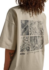 Personne portant un t-shirt beige oversize avec une impression de carte aérienne noire divisée en six panneaux et un texte incluant "ATW" au dos.