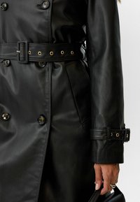 Zwarte neplederen trenchcoat met een dubbelgebreide ontwerp, een met goudaccenten versierde riem en grote knopen. Heeft een getailleerde pasvorm en gestructureerde mouwen.