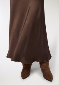 Gonna di raso marrone con una texture liscia e orlo svasato, abbinata a stivaletti marroni in camoscio con punta a punta e dettaglio a risvolto.