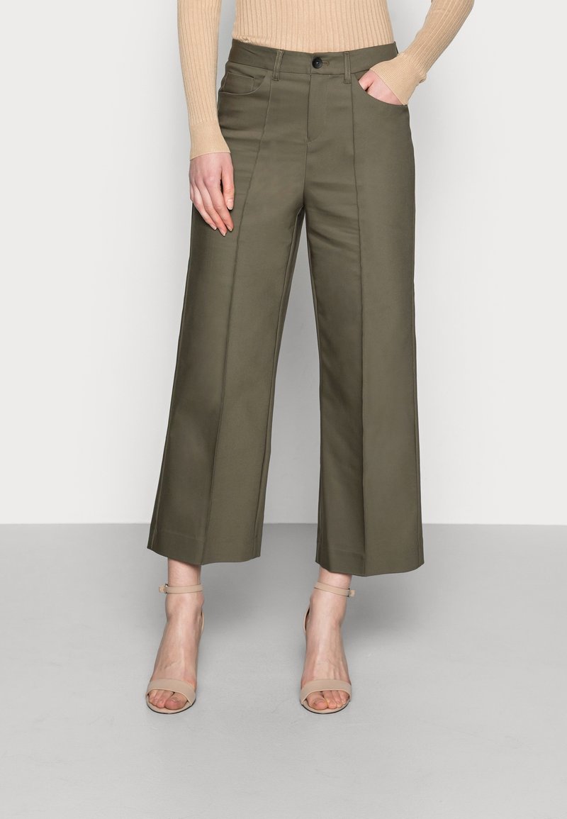 Femme portant un pantalon large court taille haute vert olive avec coutures sur le devant, un pull côtelé beige et des sandales beiges à bride cheville.