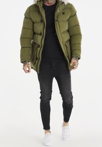 Olivengrøn pufferjakke med en hætte, der er foret med pels, med knaplukninger, to forlommer og sorte detaljer. Båret over en sort skjorte og jeans.