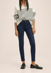 Pletená mikina v krémovo-černých pruzích s polovičním zipem, kombinovaná s tmavě modrými úzkými džíny a černými mokasínami, představující neformální outfit.