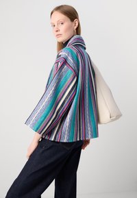 Veste rayée colorée avec un large col, des motifs complexes dans des nuances de bleu, de rose et de blanc, accompagnée de pantalons en denim noir et d'un sac crème.
