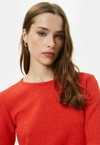 Rode, gestructureerde trui met een ribpatroon en ronde halslijn. Model heeft lang bruin haar en draagt zilveren oorbellen.