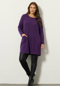 Tunika lunga viola con scollo rotondo, due tasche frontali e una vestibilità rilassata, abbinata a leggings in ecopelle nera e stivali alla caviglia.