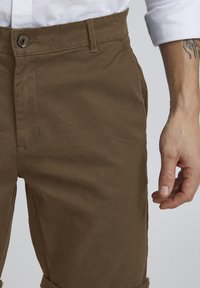 Shorts en coton marron avec une texture lisse, dotés d'une fermeture à bouton, de poches latérales et de revers. La peau est visible sur la main.