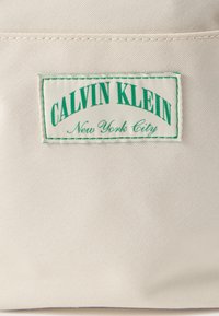 Ετικέτα Calvin Klein σε μπεζ ύφασμα, με πράσινο κείμενο, και το "New York City" σε μικρότερο, καλλιγραφικό γραμματοσειρά από κάτω. Φόντο από υφή υλικού.