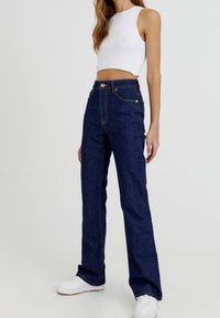 Jeans de mezclilla de tiro alto y color azul oscuro, con costuras visibles y cinco bolsillos, combinados con un top corto blanco y zapatillas blancas.