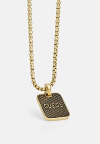 Gouden halsketting met een getextureerde rechthoekige hanger met de gegraveerde tekst "GUESS". De ketting heeft schakels en een gepolijste afwerking.
