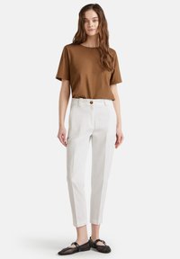 Donna con lunghi capelli castani che indossa una maglietta marrone a maniche corte, pantaloni bianchi a vita alta e scarpe basse nere con un design in rete.