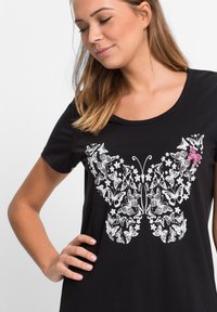T-shirt nero con un design di farfalla bianca arricchito da elementi floreali e un singolo accento rosa. Maniche corte e scollo rotondo.