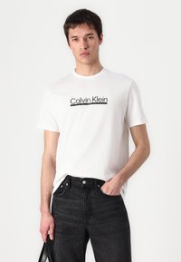 Camiseta de algodón blanca con el logo de "Calvin Klein" en negro. Combinada con jeans de mezclilla oscura, mostrando un ajuste clásico y un detalle minimalista.