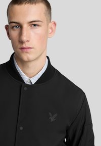 Giubbotto bomber nero con collo a coste, caratterizzato da un sottile logo dell'aquila sul petto. Indossato sopra una camicia a righe blu e bianche con colletto.