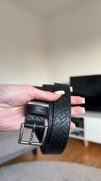 Ceinture en cuir noir avec un motif géométrique texturé, boucle en métal argenté et texture lisse. Tenue à la main sur un fond neutre.