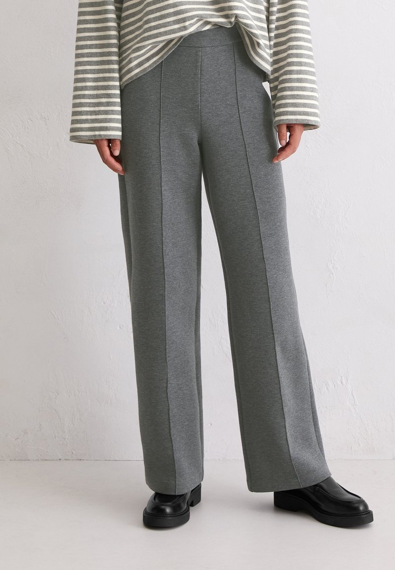 Szare szerokie spodnie wykonane z miękkiego materiału, z dodatkowymi szwami bocznymi, noszone z czarnymi butami na platformie. Zauważalna subtelna struktura heather.