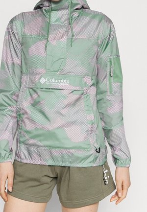 Cortavientos Columbia con camuflaje verde y lavanda, con bolsillo frontal y bolsillo en la manga con cremallera, combinado con pantalones cortos Columbia en verde oliva.