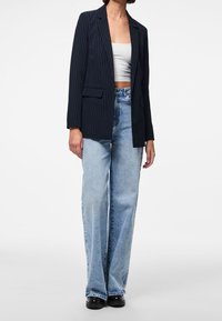 Femme portant un blazer bleu marine à fines rayures, un haut court blanc, un jean large bleu délavé et des chaussures plateformes noires sur un fond uni.