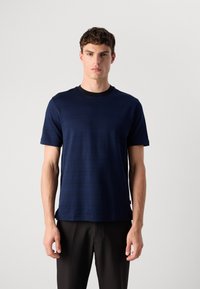 Jovem em pé, vestindo uma camisa de manga curta azul escura com listras horizontais subtis e calças pretas, contra um fundo cinza liso.