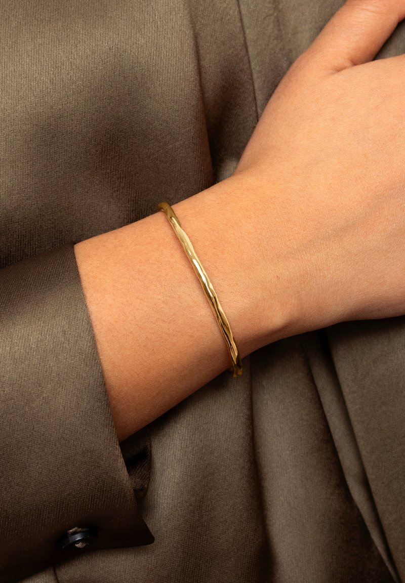Main portant un bracelet jonc fin et texturé en or sur un vêtement en tissu soyeux marron.