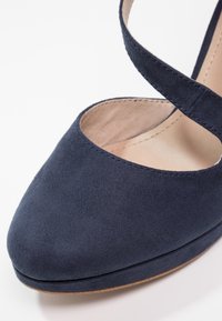 Hauteur en daim bleu marine foncé avec un bout rond et une sangle de cheville croisée. Texture lisse avec un léger éclat et semelle intérieure beige.