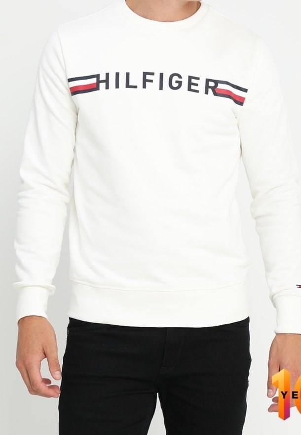 Sweat-shirt blanc à manches longues avec le texte « HILFIGER » et des rayures rouges, blanches et bleues sur la poitrine, porté avec un pantalon noir.