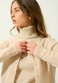 Jersey de cuello alto de color beige con costillas, combinado con un cárdigan beige con botones. Suave textura, mangas largas y colores naturales crean una apariencia acogedora.