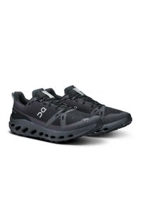 Chaussures de sport noires avec une tige en mesh texturé, au design épuré, et une semelle en caoutchouc présentant des découpes circulaires pour une meilleure adhérence. Résistantes à l'eau.