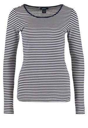 Femme portant un haut à manches longues rayé noir et blanc, pantalon beige, bottines noires, tenant un sac bandoulière noir et beige.