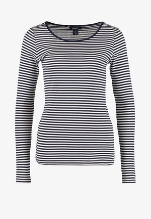 Frau trägt ein schwarz-weiß gestreiftes Langarmshirt, beige Hose, schwarze Knöchelstiefel, hält eine schwarz-beige Umhängetasche.
