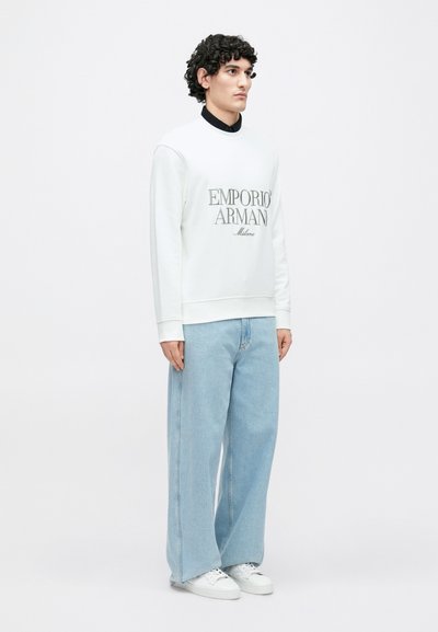 Sweatshirt branca com o logo "Emporio Armani", combinada com jeans denim oversize azul-claro e ténis brancos. Design simples e clean.
