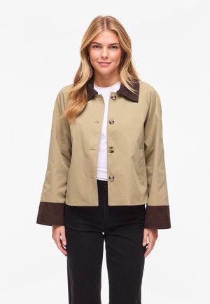 Blondīne, kas valkā bēšu jaku ar tumši brūnu apkakli un aprocēm, baltu kreklu un melnas bikses, stāv un raugās uz priekšu.