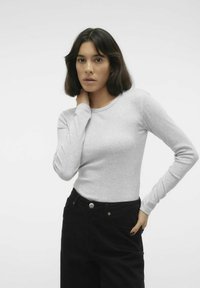 Maglietta grigia a maniche lunghe con scollo rotondo, abbinata a pantaloni neri a vita alta con tasca frontale e dettagli in metallo visibili.