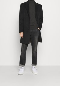 Cappotto nero lungo sopra un dolcevita grigio scuro, abbinato a jeans scuri e sneakers bianche, con orli arrotolati e dettagli discreti nelle tasche.