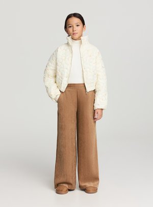 Giacca trapuntata color crema con motivo floreale, collo alto, dolcevita bianco, pantaloni marroni a gamba larga in velluto a coste e scarpe slip-on color cuoio.