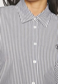 Donna che indossa una camicia a righe verticali bianco e nero con colletto a punta e logo ricamato sul petto.